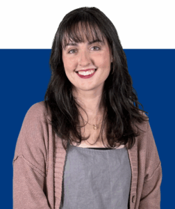 Anna Hamrick, with long dark hair and bangs, smiles in a mauve cardigan and gray top against a blue and black background.